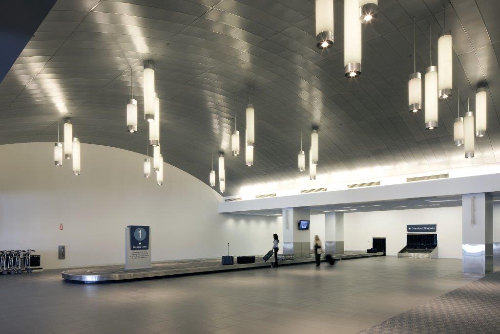 Two passengers at the baggage claim carousel. Pendant lights are hanging from the aluminum ceiling