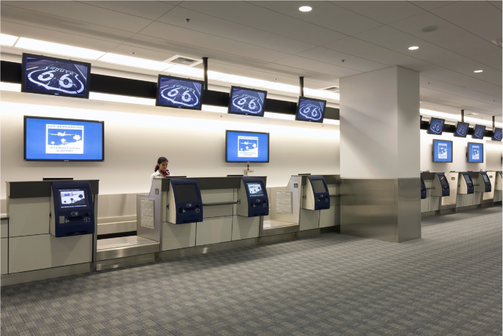 Ticketing agent stands at ticketing counter. Monitors are hanging above and behind the counter.